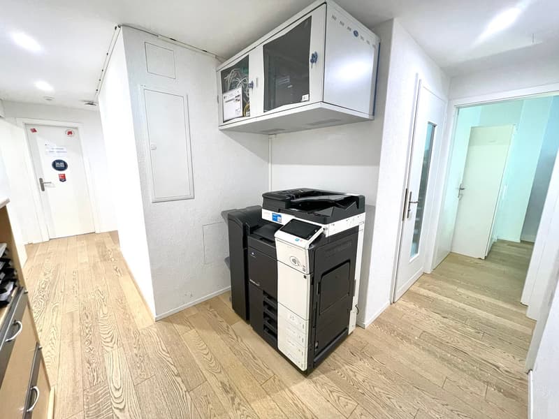 The image shows an office space with a printer/copier machine and a microwave oven mounted on the wall. The room has a hardwood floor and white walls, and there are doors leading to other rooms or areas.