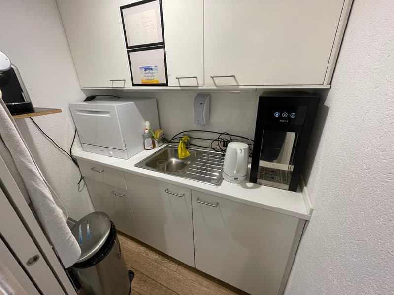Compact kitchen with white cabinets, stainless steel sink, dishwasher, and washing machine. There is also a water dispenser and some small appliances on the counter.