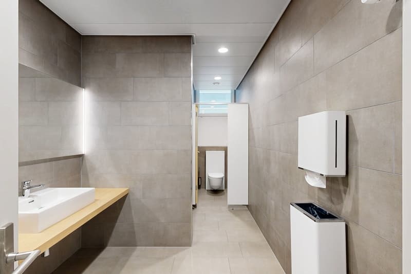 clean bathroom with white sink, silver faucet, white trash bin, and grey tiled walls