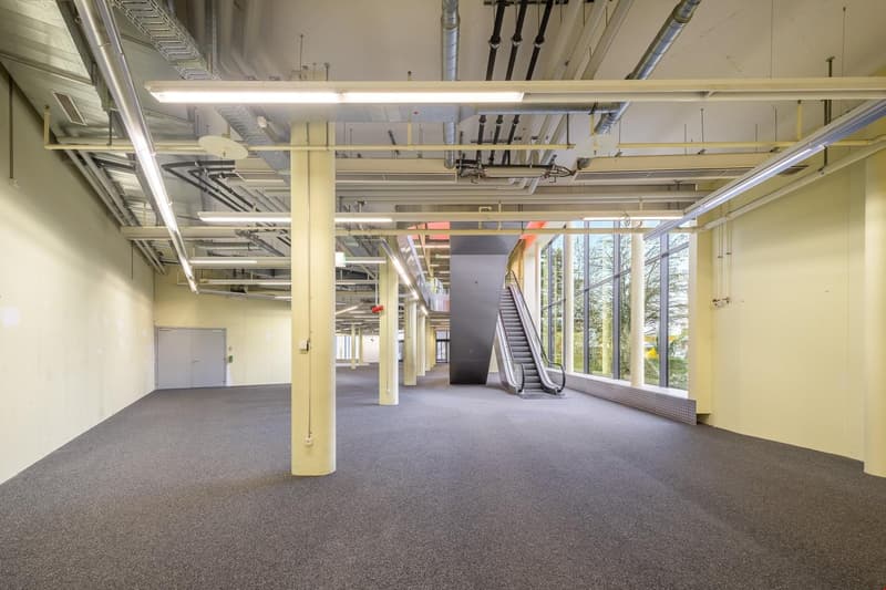 wide empty corridor, fluorescent lights, central ceiling columns, white walls, double glass windows, and a closed door on the left