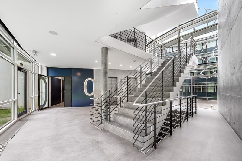 Modern and minimalist interior design with concrete floors, metal staircase with geometric patterns, and large windows providing natural light. The space features a sleek and industrial aesthetic with a focus on clean lines and open layout.