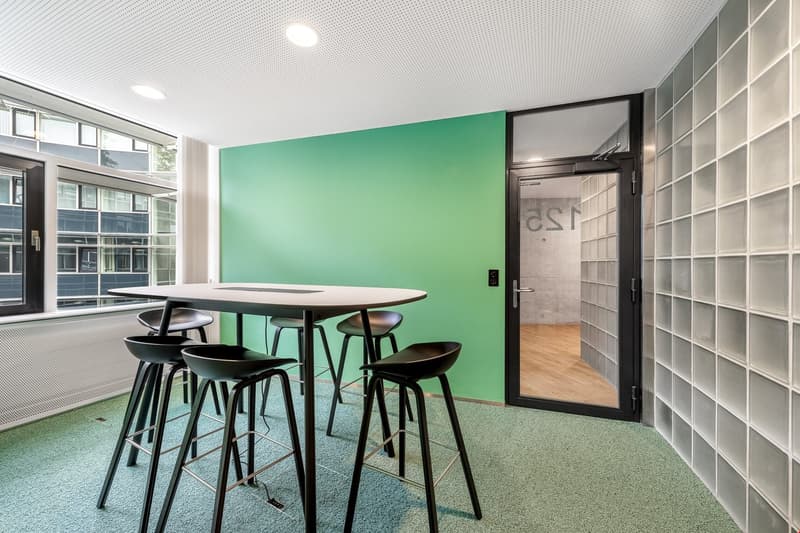 Modern office space with green accent wall, round table with black stools, glass windows, and glass block wall