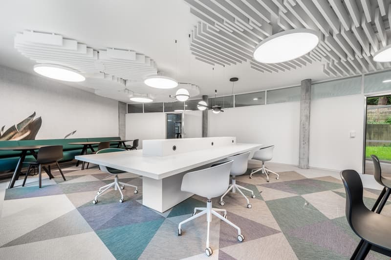 Modern and open office space with white desks, swivel chairs, and patterned carpet flooring. The ceiling features recessed lighting and a geometric design. The space also includes a glass partition wall and a view of the outdoors through a window.