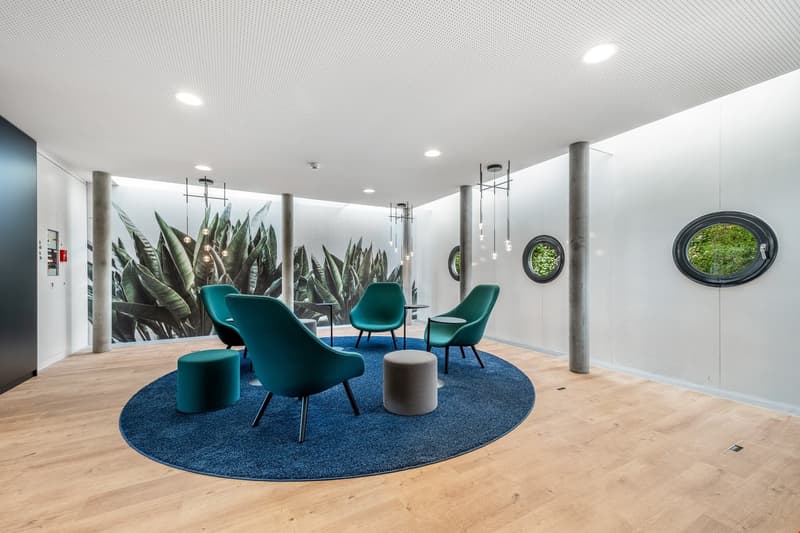 Spacious office lounge with large wall mural of tropical plants, teal armchairs and ottomans, round blue rug, wooden flooring, and circular wall mirrors providing a view of the outdoors.