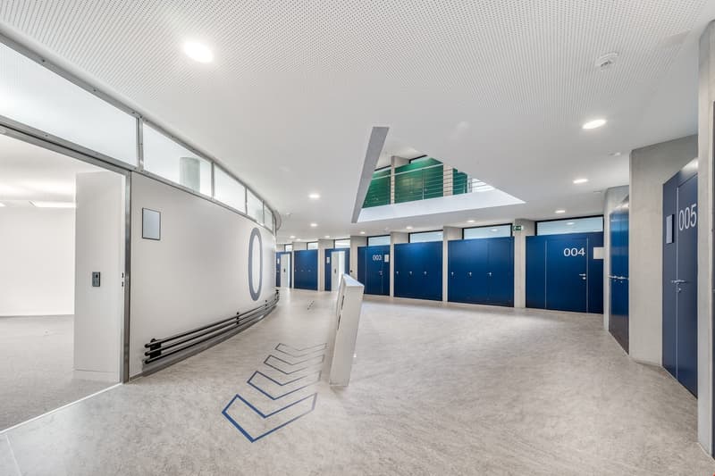 The image shows a long, well-lit hallway with a high ceiling and a glass wall on one side. The hallway has numbered lockers or storage units on both sides, with blue doors. The floor is a light-colored tile or concrete. The hallway appears to be part of a