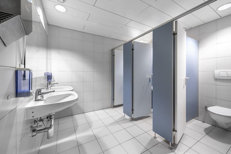 Public bathroom with white tiles, two sinks, three stalls, and a toilet.
