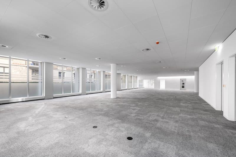 Empty room with white walls, gray carpet, large windows, and ceiling with air vents