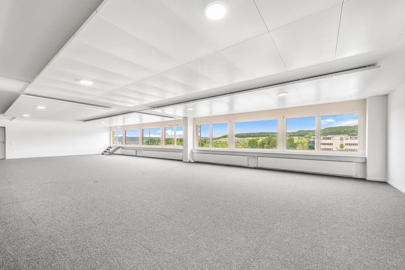 An empty room with gray carpeting, a white ceiling, and large windows offering a scenic view