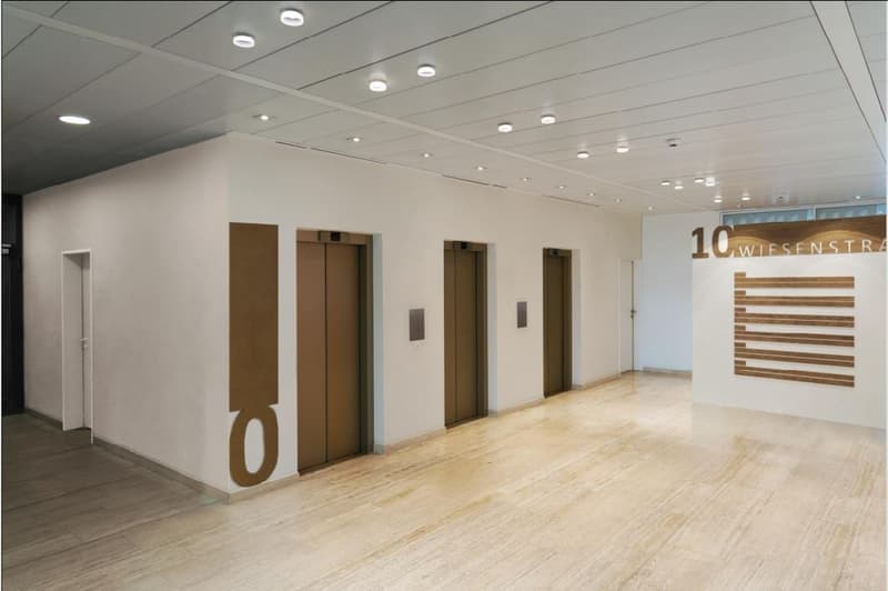 Empty hallway with three elevators, wooden floor, white walls, ceiling lights