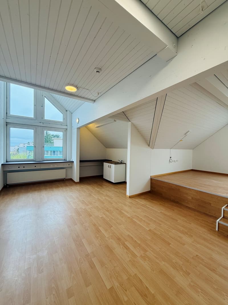 Empty room with wooden floor, white walls, large windows, small kitchen area, and a small raised platform.