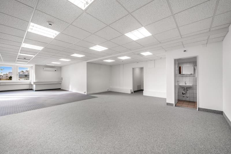 Empty room with white walls, gray carpet, ceiling lights, and a window with a view outside.