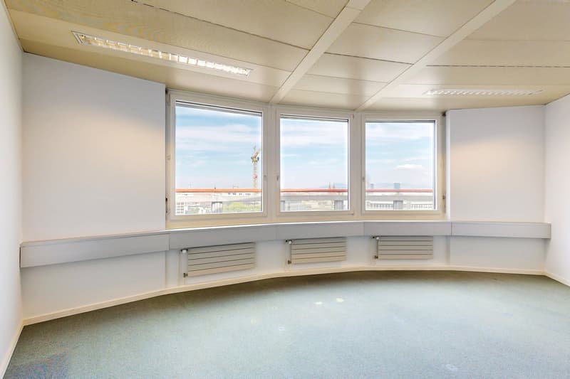 Large open office space with floor-to-ceiling windows providing a view of the city skyline. The room has a suspended ceiling with lighting fixtures and built-in storage units along the walls.