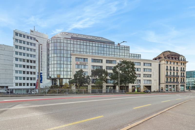 The image shows a modern glass and steel office building with a curved roof, surrounded by older, more traditional-style buildings. The building appears to be the Medizinischen Universität building, as indicated by the sign on the facade. The street in fr