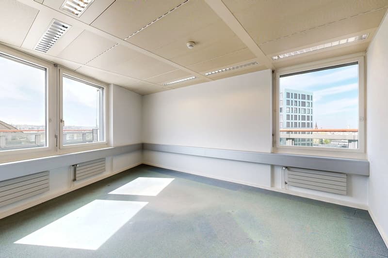 The image shows a large, open office space with white walls, a tiled floor, and large windows providing natural light. The room has a suspended ceiling with recessed lighting fixtures. There are built-in storage units or shelves along the walls.