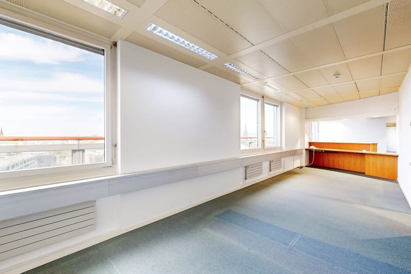 Large open office space with white walls, suspended ceiling with lighting, and a wooden reception desk. The room has large windows providing natural light and a view of the surrounding area.