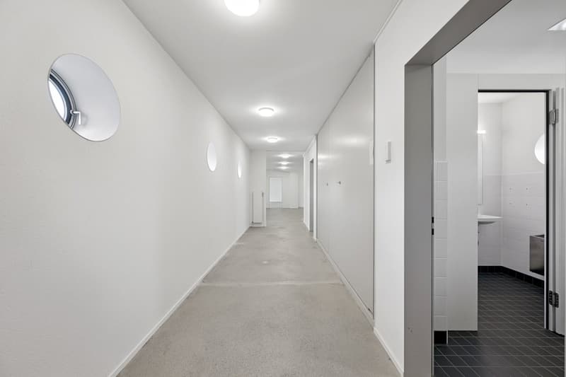 long hallway, white walls, circle window, gray tile floor, square ceiling lights, bathroom door