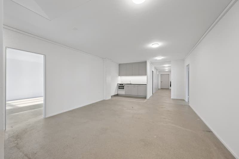 An empty room with white walls and beige flooring, kitchen with cabinets and countertop, oven, and sink.