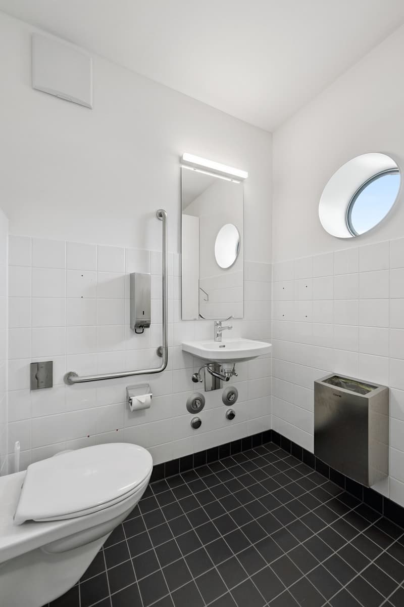 A toilet, sink, mirror, and a tissue dispenser, all with a modern and minimalist design. There is also a round window on the wall.