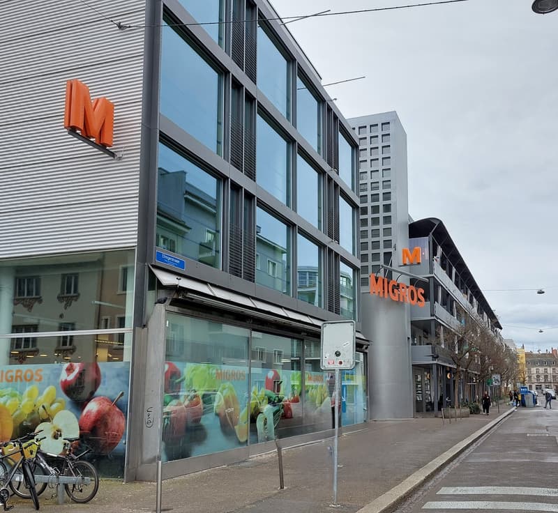 Modern building with large glass windows, large sign reading 'Migros'. Bicycles parked to the left.