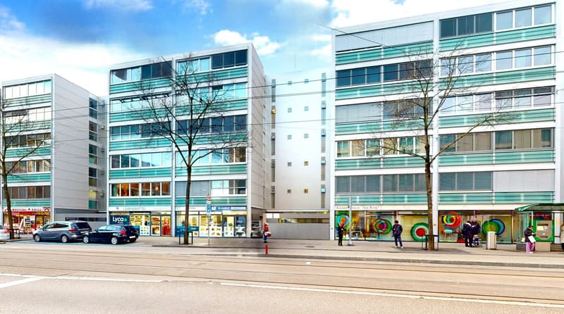 Three buildings with large windows, on a street, with people walking, cars parked, trees, and shops in front.