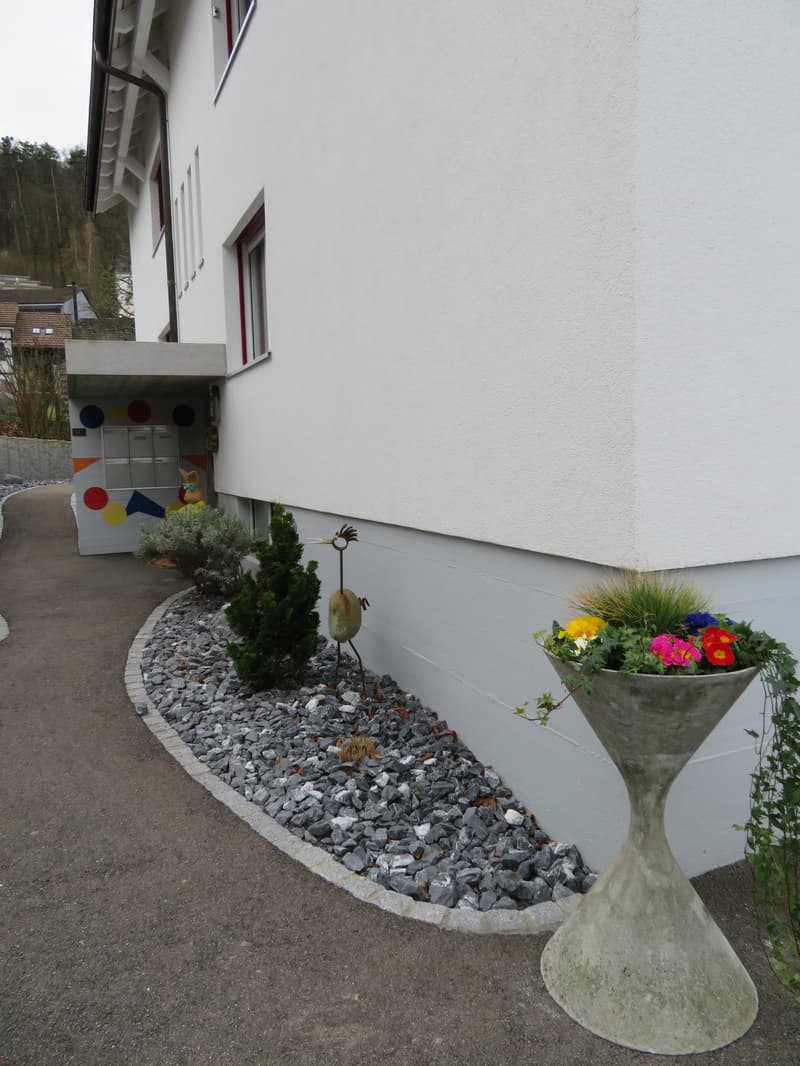 concrete gravel path, two green bushes, colorful flower pot, decorative metal piece, white walls