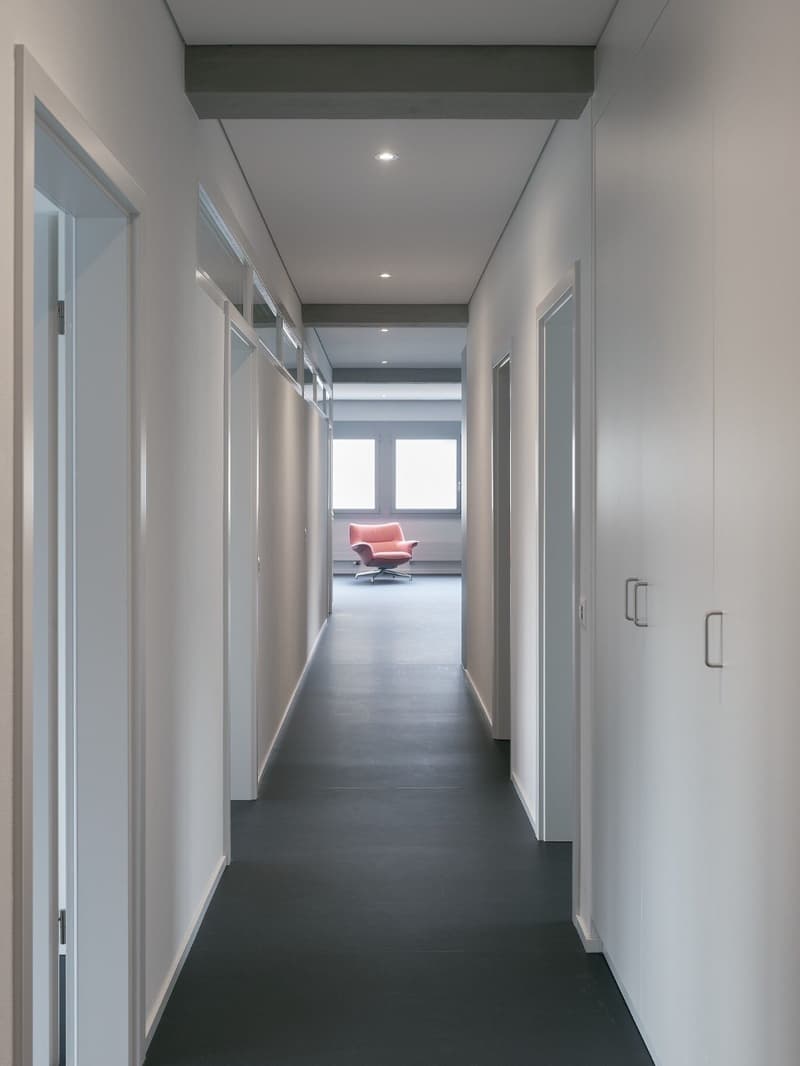 Long, narrow hallway with white walls, dark hardwood floors, recessed lighting, and a glass-paneled door leading to a room with a pink chair visible at the end.