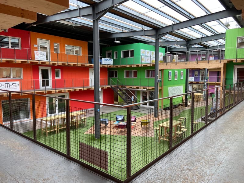 multicolored living units with wooden stairs, central playground with grass, multiple seating areas, metal fences, central walkway, clear glass roof, multiple balconies