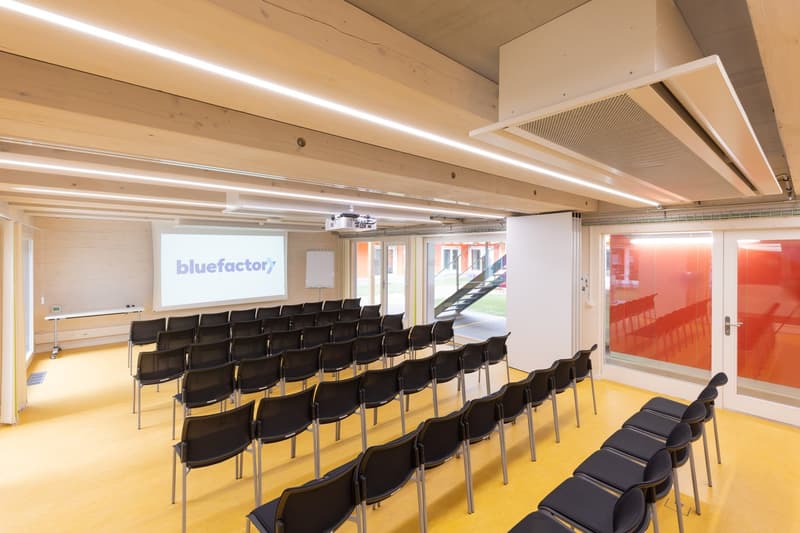 Yellow flooring, ceiling with exposed beams, chairs arranged in rows, whiteboard, projector screen, glass doors