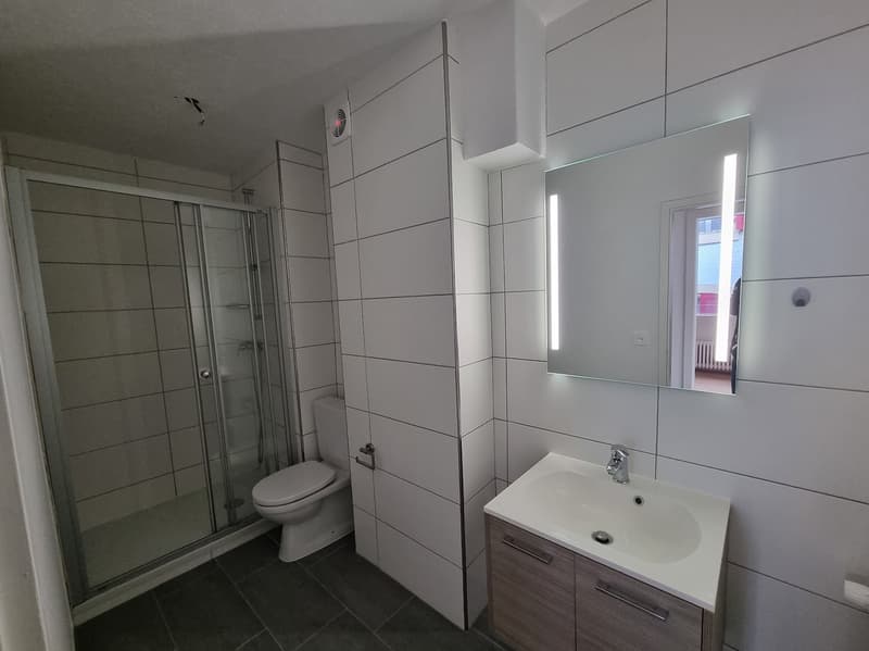A white bathroom with a white sink, silver faucet, and mirror, and a shower cubicle, white tiled walls and floor, a white toilet, and a window behind the mirror.