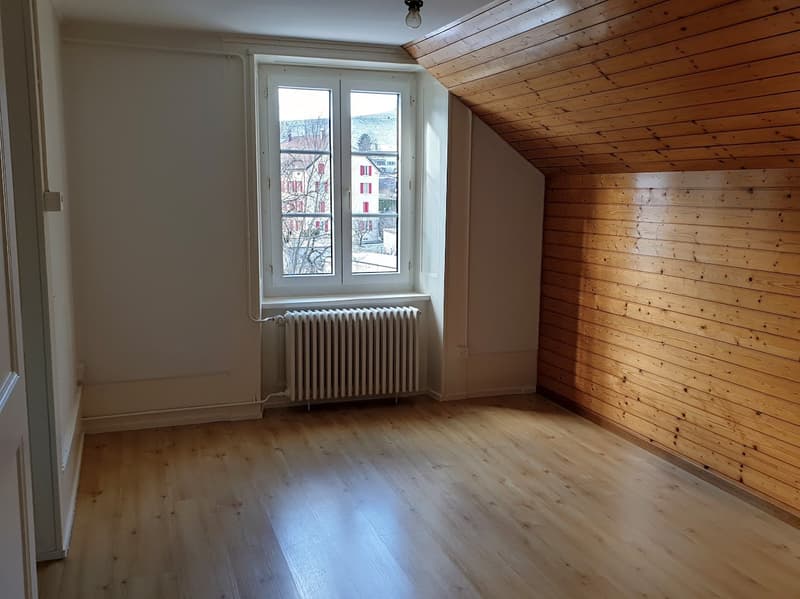 This image shows a room with a slanted wooden ceiling and white walls. The room has large windows that provide natural light and a view of the surrounding buildings. The floor is made of light-colored hardwood. There is a radiator visible under the window