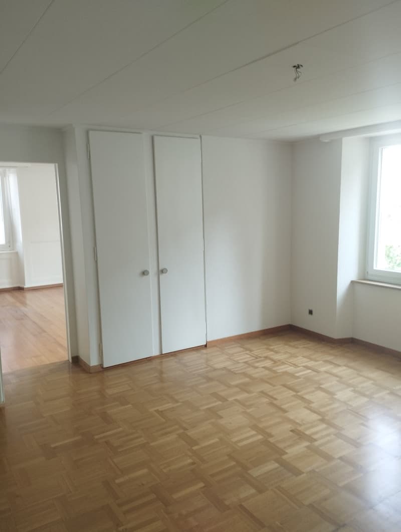 This image shows an empty room with white walls and a hardwood floor in a herringbone pattern. The room has two white closet doors and a window, indicating it could be a bedroom or living space. The room appears to be in a clean, minimalist style with no