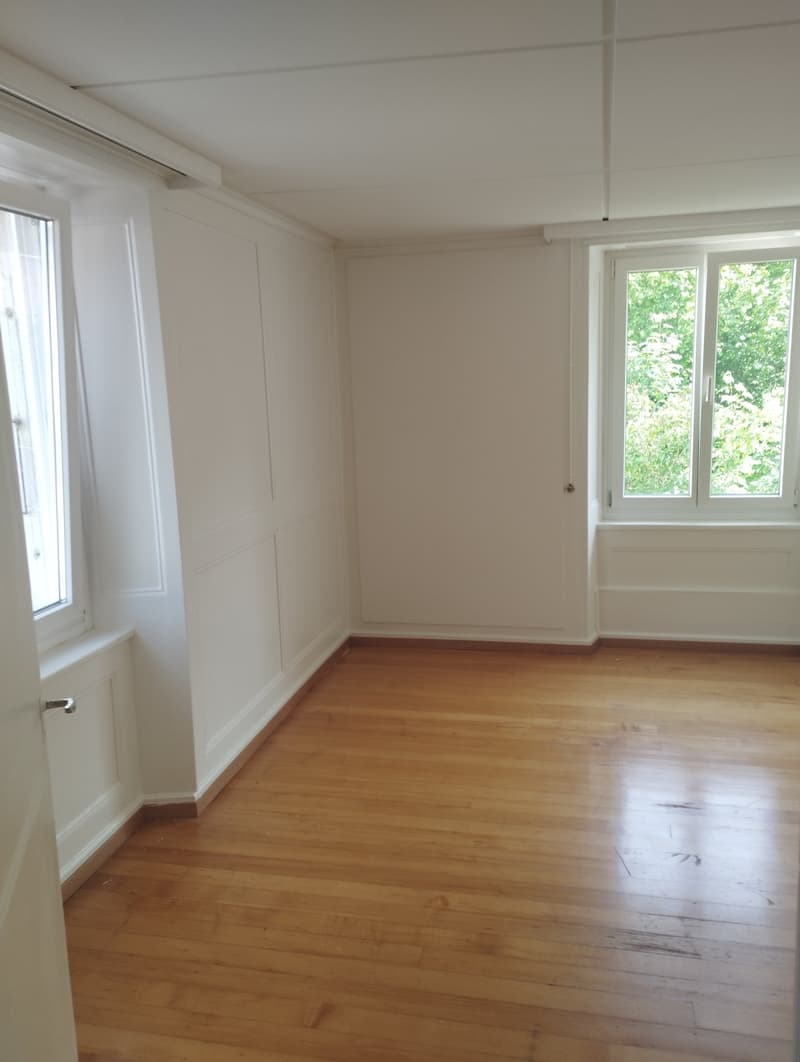 Spacious bedroom with hardwood floors, white walls, and large windows overlooking greenery outside.