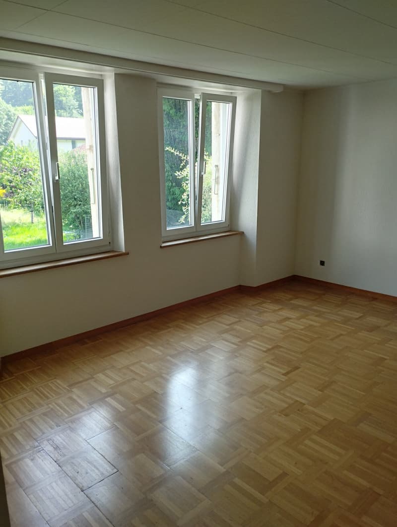 This image shows a spacious room with large windows overlooking a garden. The room has a hardwood floor in a herringbone pattern and white walls. The room appears to be empty, ready for furnishing and decorating.