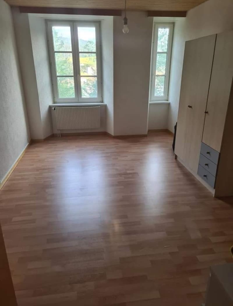 Room with wooden floors, white walls, two windows, white radiators, wooden cabinet with drawers.