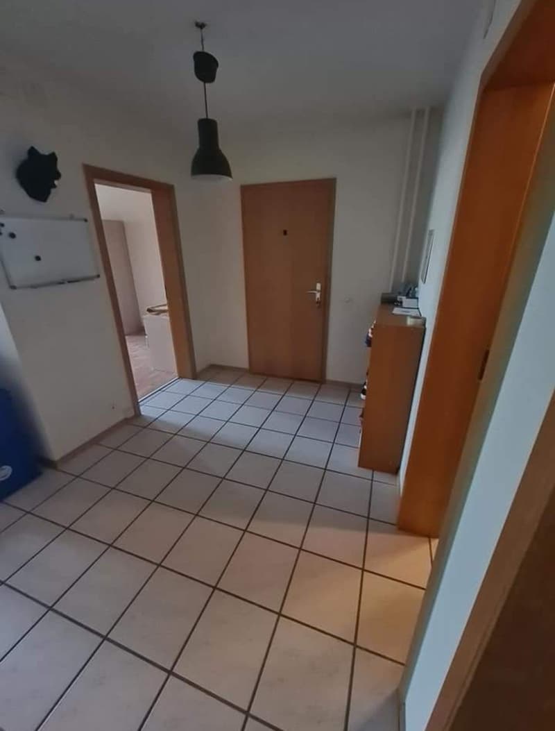 Empty hallway with white tile floor, white walls, wooden doors, mounted whiteboard, framed picture, cabinet with items
