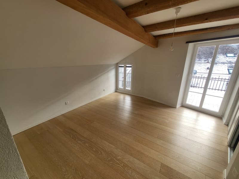 Empty room, wooden floor, white walls, wooden ceiling beams, two windows, balcony with railings