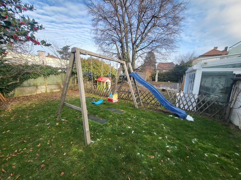 Grassy lawn with a tree and hedge, wooden play equipment including a swing set, slide, and small house, wooden fence, house walls in the background, and cloudy sky.