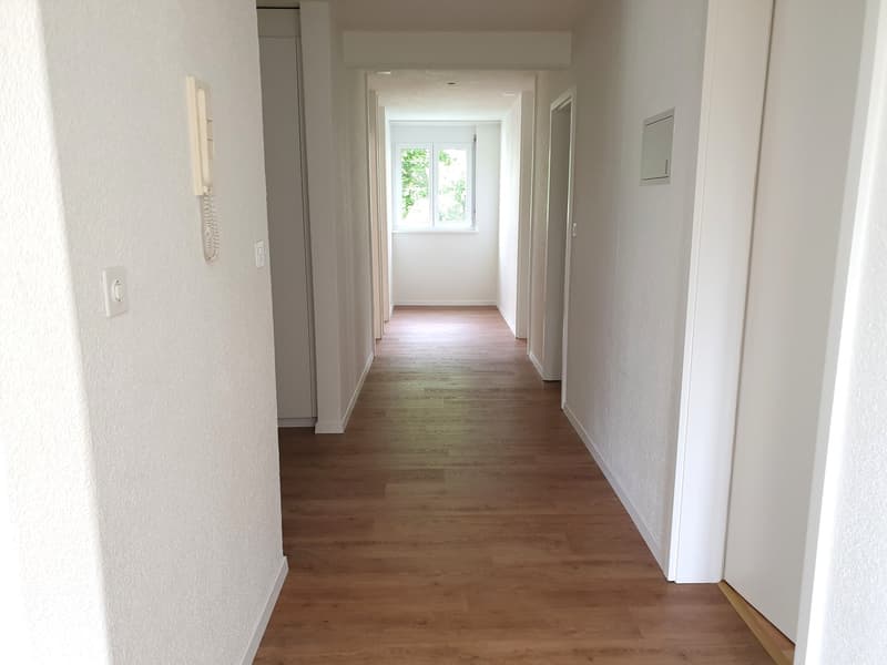 Empty hallway with wooden floor, white walls, and a window at the end.