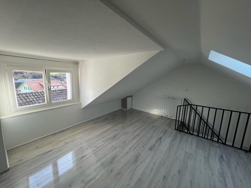 Empty attic room, white walls, wooden floors, a window with an outside view, slanted ceilings, and a handrail staircase.