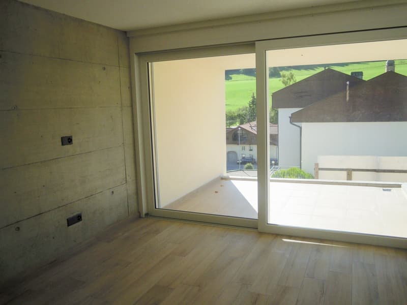 Empty room with wooden floor, large glass sliding doors leading to balcony, concrete walls, green field view outside