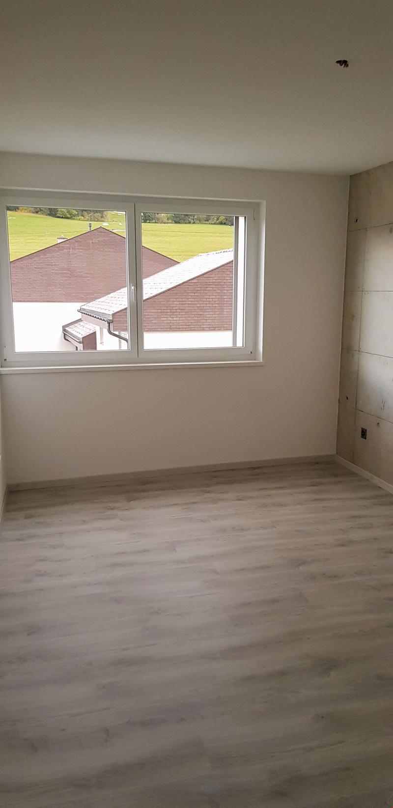 An empty room with white walls, wooden floor, a large window, and a power outlet on the right wall