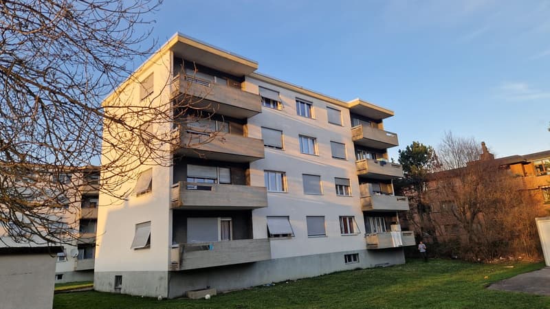 Concrete building with balconies, 5 stories, multiple windows, located on a grassy area with other buildings in the background