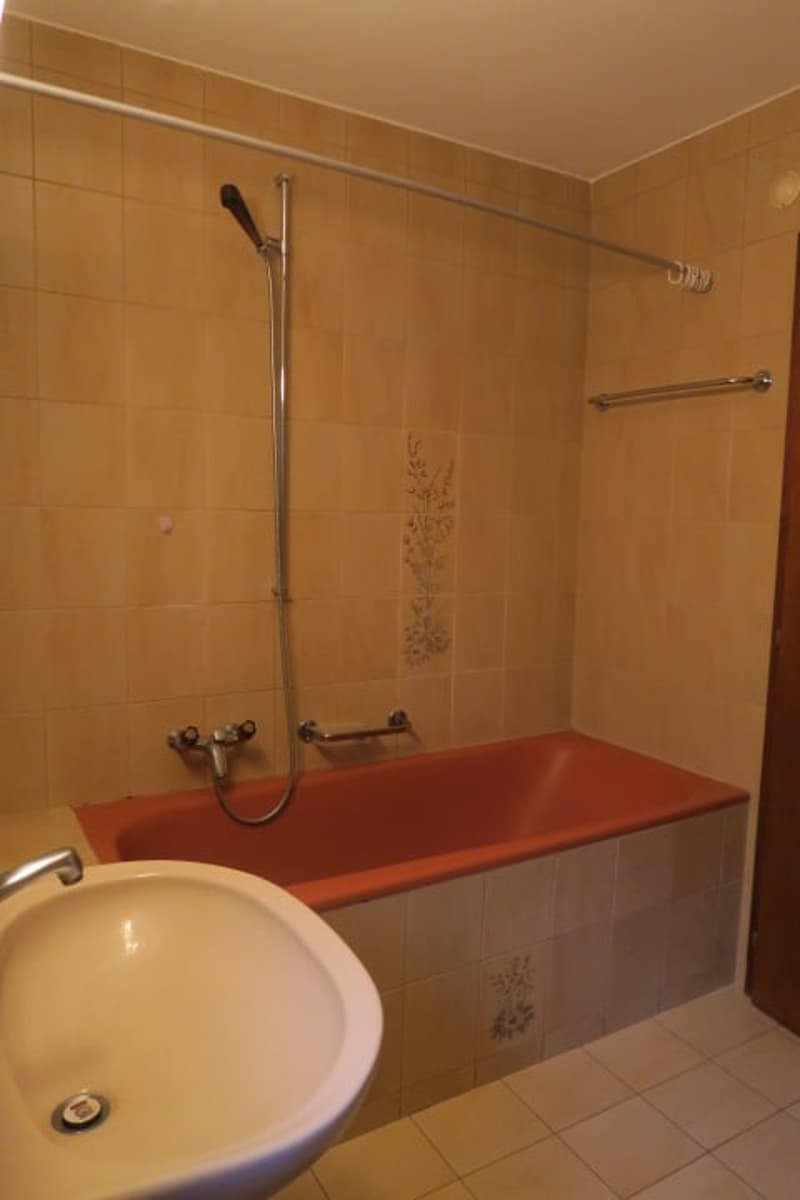 Bathroom with bathtub and sink. Bathtub is orange with tiled walls. Tiled floors and walls. Shower head and hose. Sink is white with silver faucet.