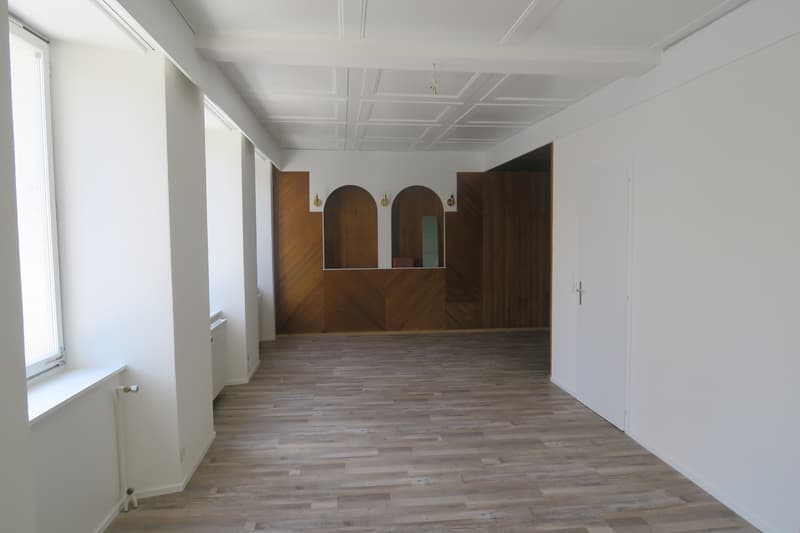 Empty room with wooden floor, white walls, wooden paneling, windows, radiators