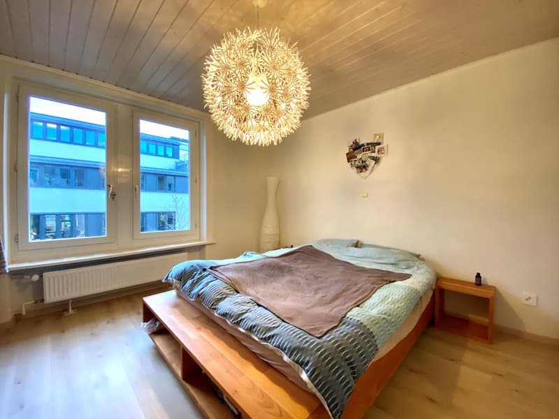 Spacious bedroom with large windows, hardwood floor, a wooden bed frame, and a decorative light fixture hanging from the ceiling.