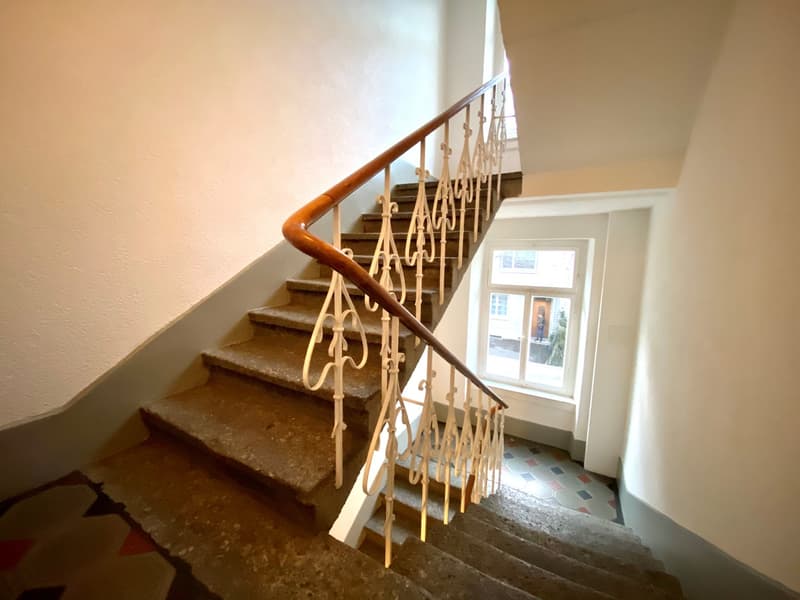 Wooden staircase with ornate metal railing, leading to a second floor or upper level of the building. The stairs appear to be made of stone or concrete, with a tiled floor visible at the bottom.