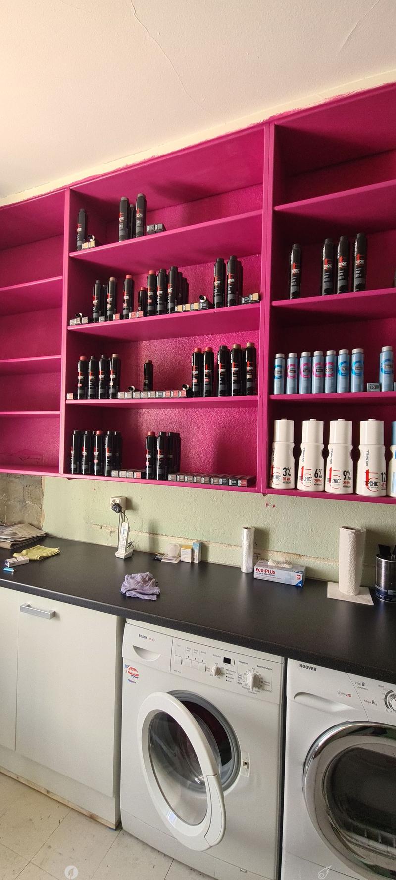 Laundry room with washer and dryer, pink shelves, bottles, and rolls on top, kitchen items, towels on floor, black countertop