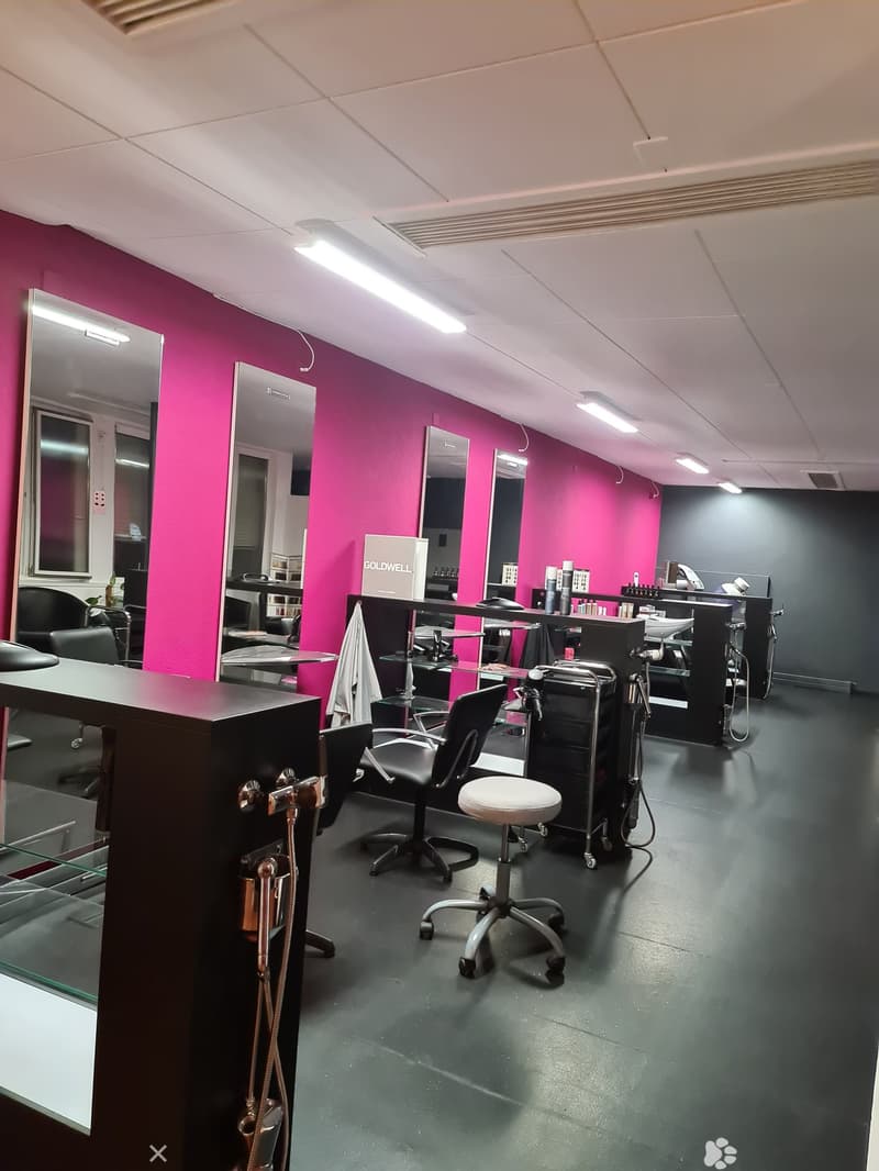 The image shows a salon with multiple black chairs, tables, and mirrors. Pink walls are visible in the background, and large glass mirrors line the room. Hair dryers, bottles, and sinks are visible at the sinks.