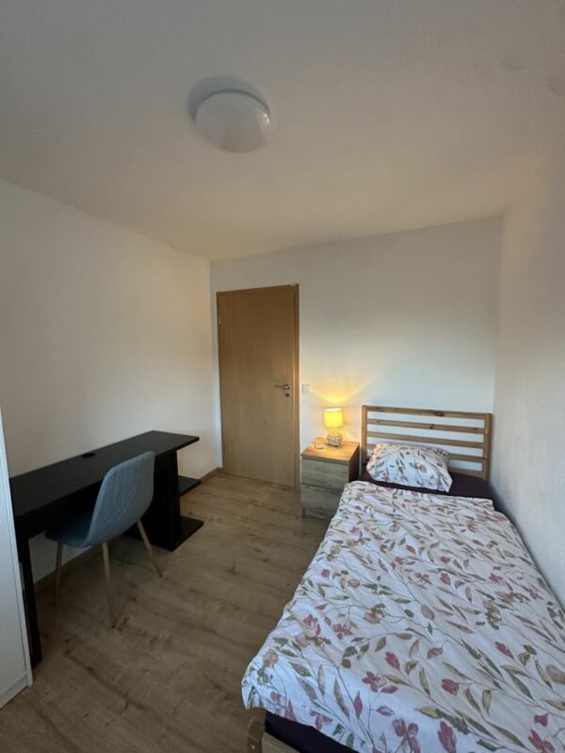 small bedroom with bed, desk, chair, nightstand, lamp, wooden floor, white walls