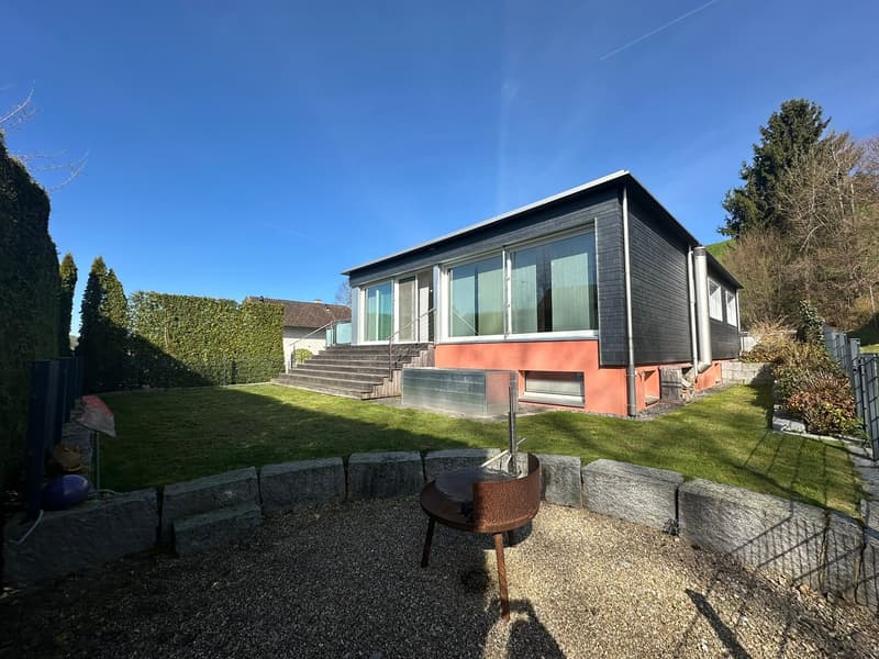Modern house with glass windows, black and orange walls, stairs leading to the entrance, a small garden with a barbecue, and a gravel pathway.